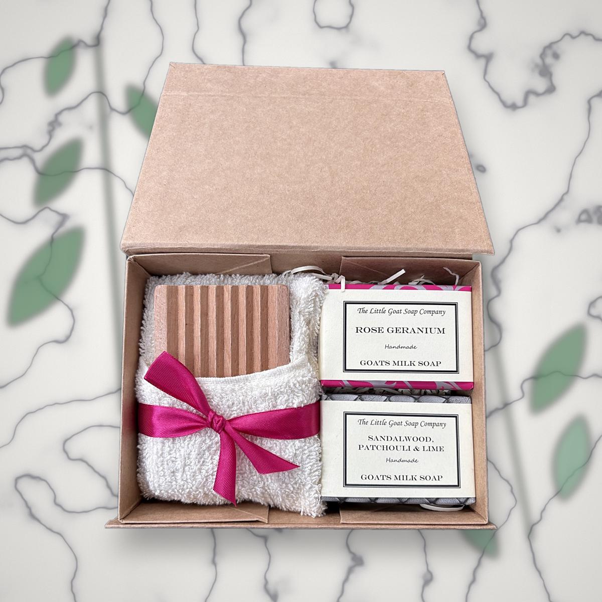 Open Little Goat Soap Company gift box with a wooden soap dish, white flannel tied with a pink ribbon, and Rose Geranium and Sandalwood, Patchouli & Lime goat milk soap bars, on a marble background with greenery