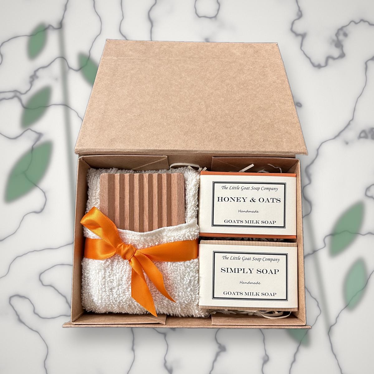 Open Little Goat Soap Company gift box with a wooden soap dish, white flannel tied with an orange ribbon, and Honey & Oats and Simply Soap goat milk soap bars, on a marble background with greenery