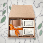 Open Little Goat Soap Company gift box with a wooden soap dish, white flannel tied with an orange ribbon, and Honey & Oats and Simply Soap goat milk soap bars, on a marble background with greenery