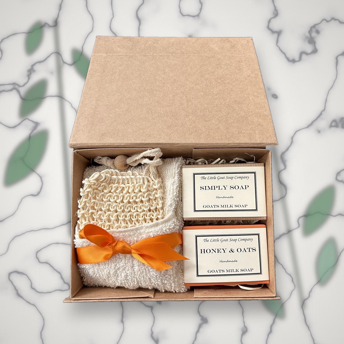 Open Little Goat Soap Company gift box with Simply Soap and Honey & Oats goat milk soap bars, a sisal soap bag and white flannel tied with an orange ribbon, on a marble background with greenery