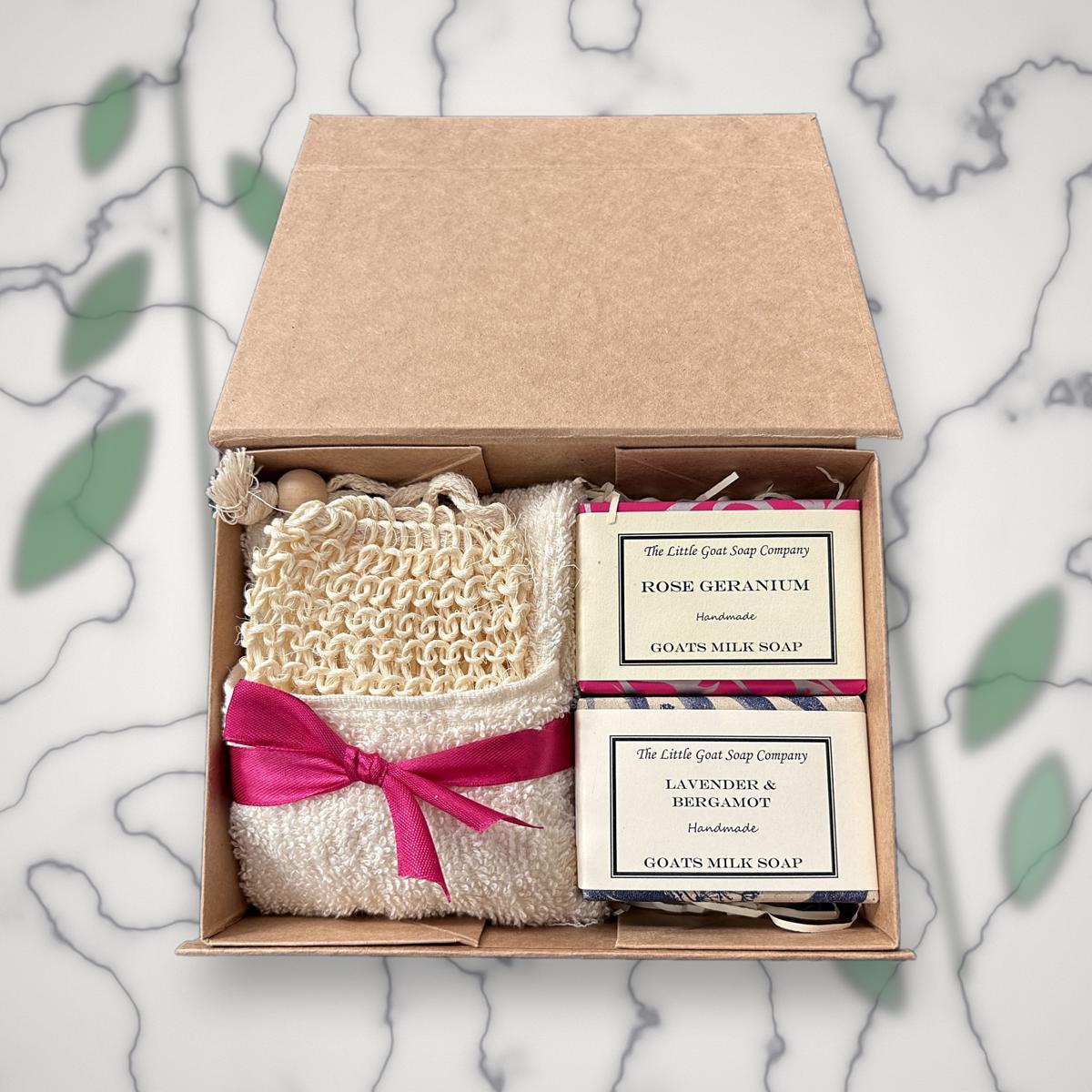 Open Little Goat Soap Company gift box with Rose Geranium and Lavender & Bergamot goat milk soap bars, a sisal soap bag and white flannel tied with a pink ribbon, on a marble background with greenery