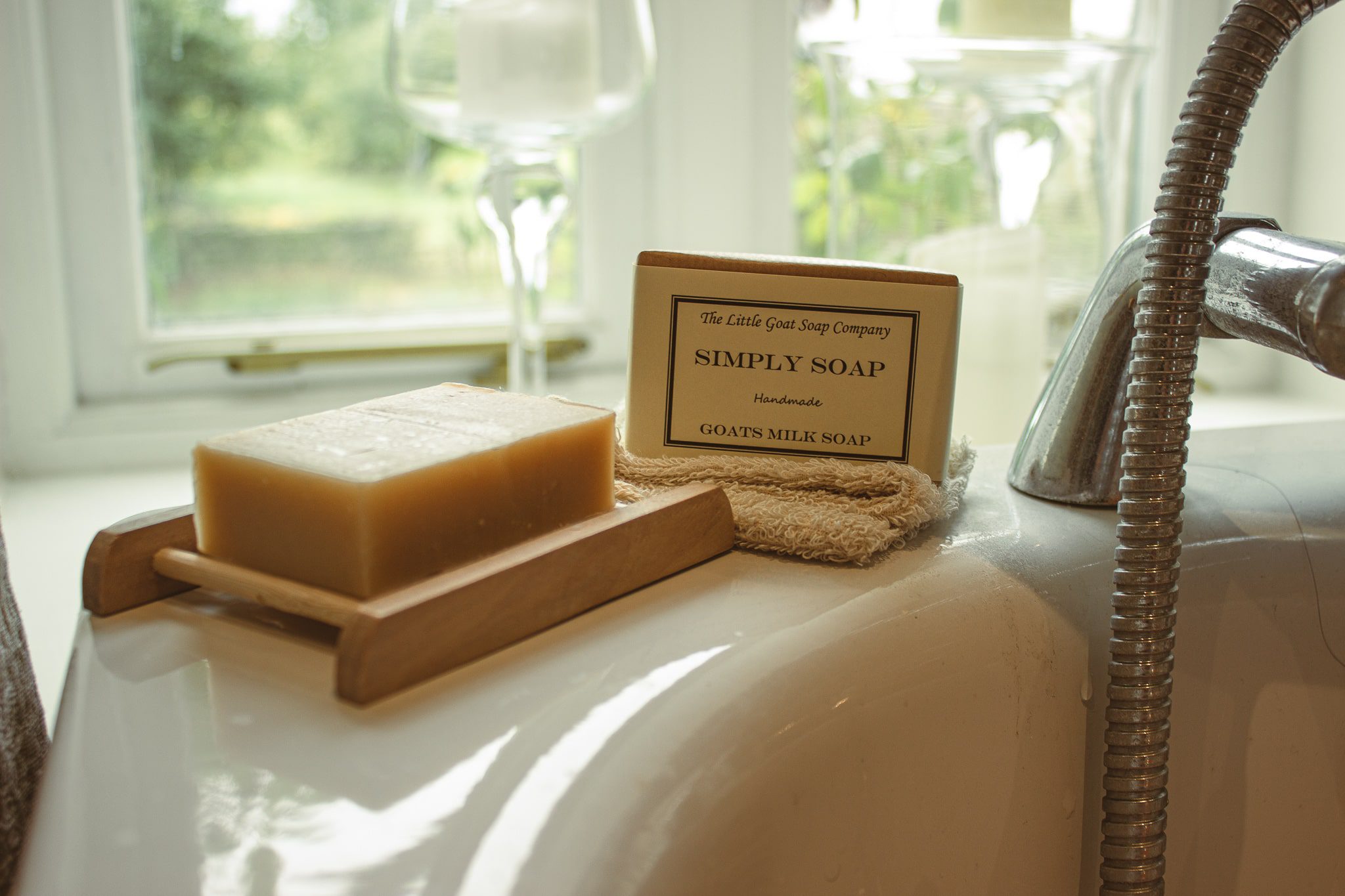 goat milk soap, on a soap tray by the bath. There is also a soap bar still in its packaging resting on a soap bag.